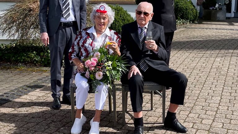 Ein älteres Ehepaar sitzt schick gekleidet in der Sonne und lächelt in die Kamera.