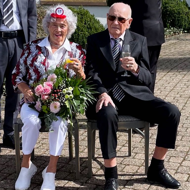 Ein älteres Ehepaar sitzt schick gekleidet in der Sonne und lächelt in die Kamera.