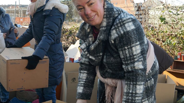 Ältere Frau in einer grau-weißen Jacke packt zwei Kartons mit Kleidung. 