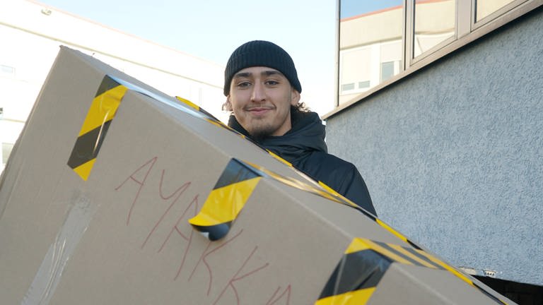 Junger Mann, mit Mütze trägt einen riesigen Karton. 