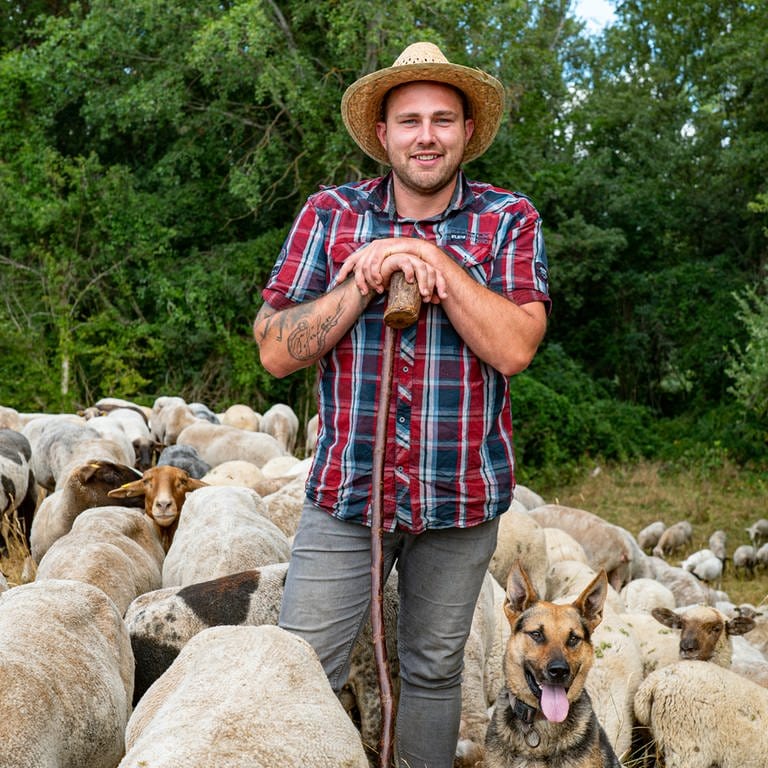 Junger Mann mit kariertem Hemd und Strohhut lehnt vor einem Wald auf einem Stock. Um ihm herum sind Schafe. 