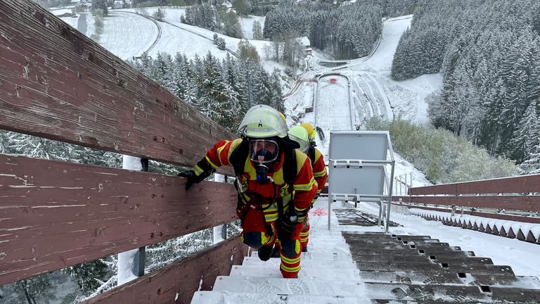 Zwei Feuerwehrleute laufen in voller Montur die verschneite Langenwaldschanze in Schonach hoch.